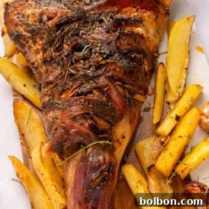 The Ultimate Roasted Lamb Leg 12 A roasted leg of lamb garnished with herbs is served on a wooden board alongside golden potato wedges and roasted garlic cloves.
