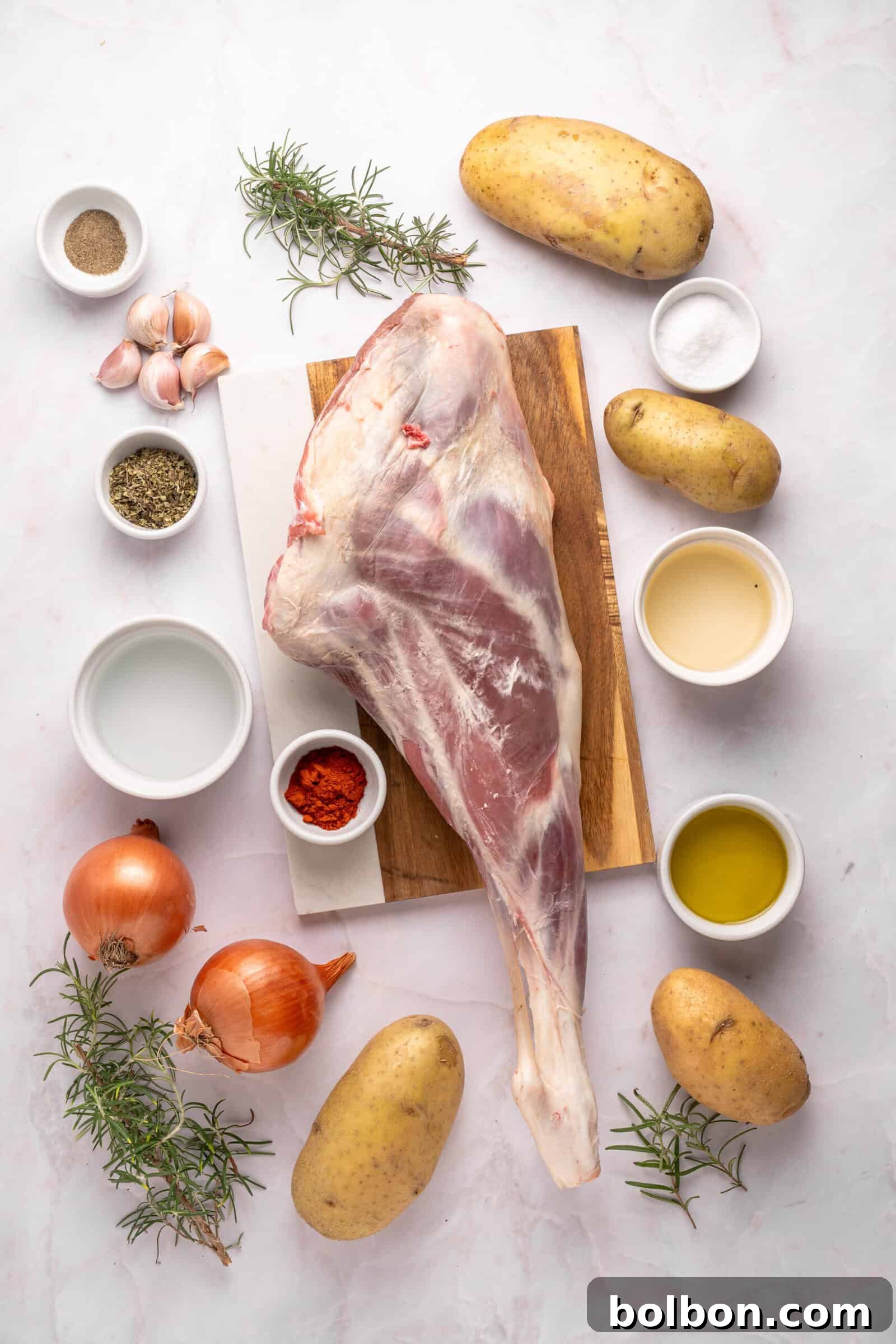 The Ultimate Roasted Lamb Leg 3 Raw lamb leg on a wooden board surrounded by potatoes, onions, garlic, rosemary, spices, salt, pepper, olive oil, and other seasonings in small bowls on a white background.