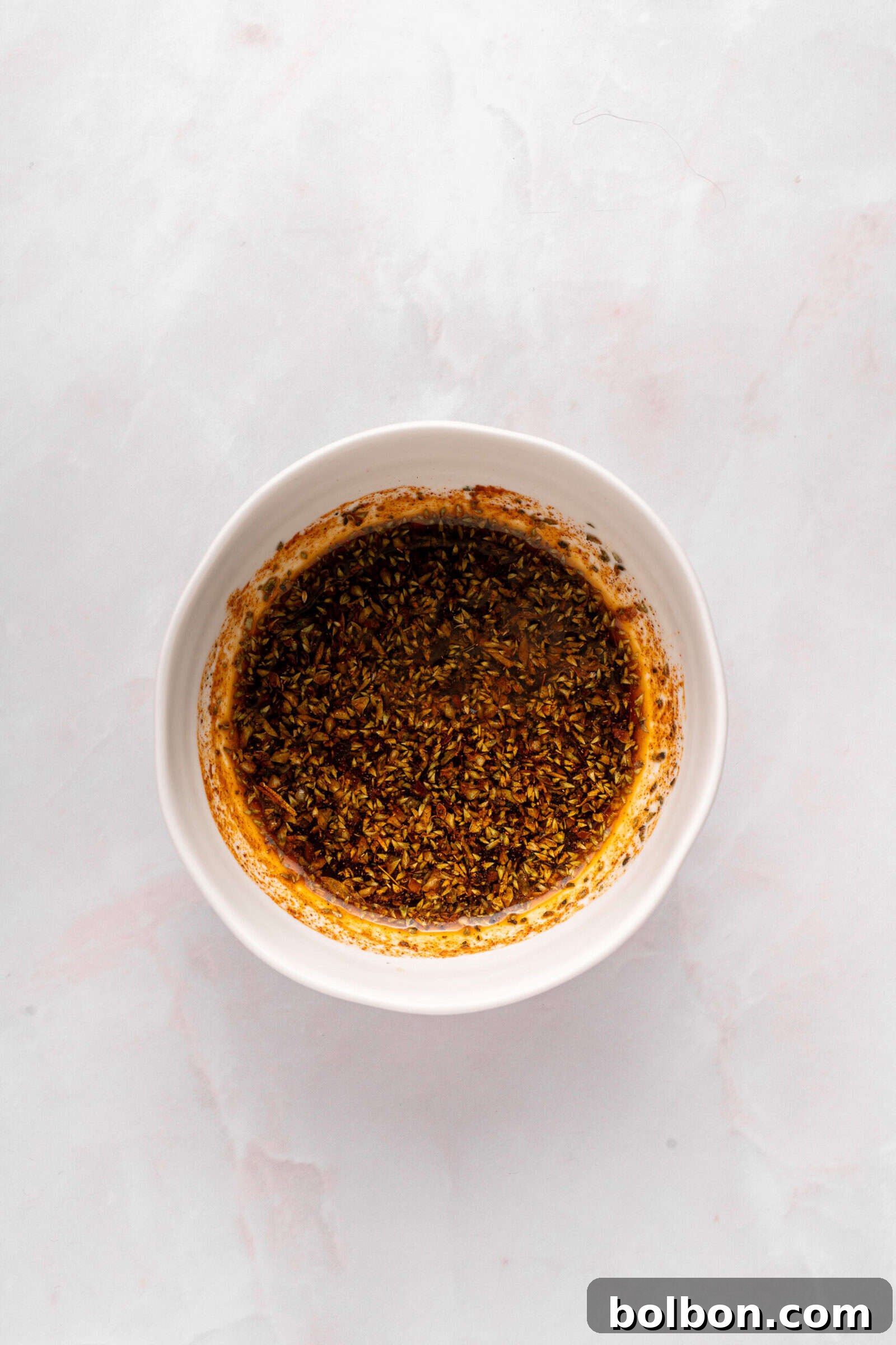 The Ultimate Roasted Lamb Leg 5 A white bowl filled with a dark, oily mixture of herbs and spices sits on a light marble surface.
