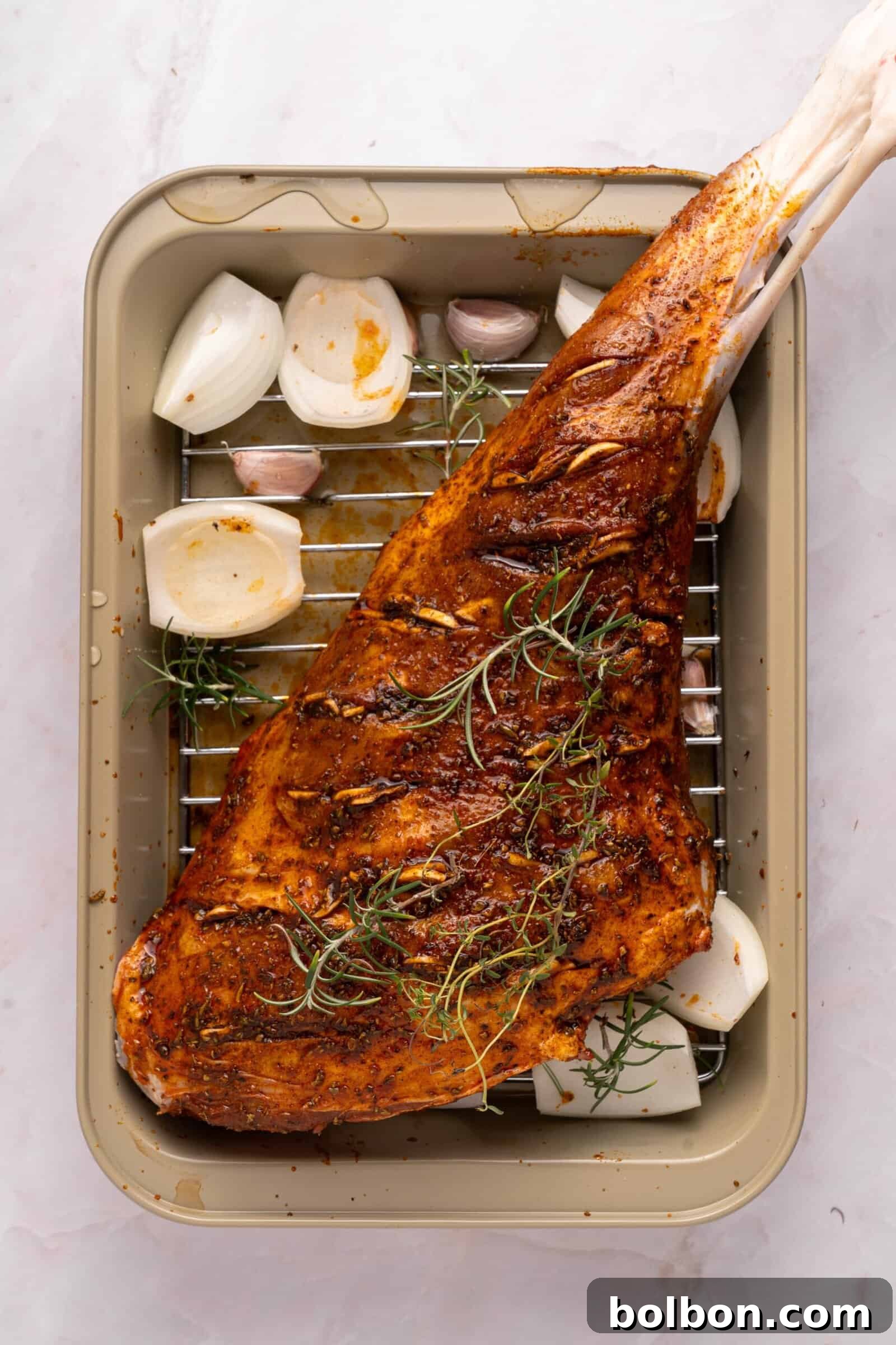 The Ultimate Roasted Lamb Leg 7 A seasoned and spiced leg of lamb sits on a rack in a roasting pan, surrounded by halved onions, garlic cloves, and sprigs of rosemary, ready to be cooked.