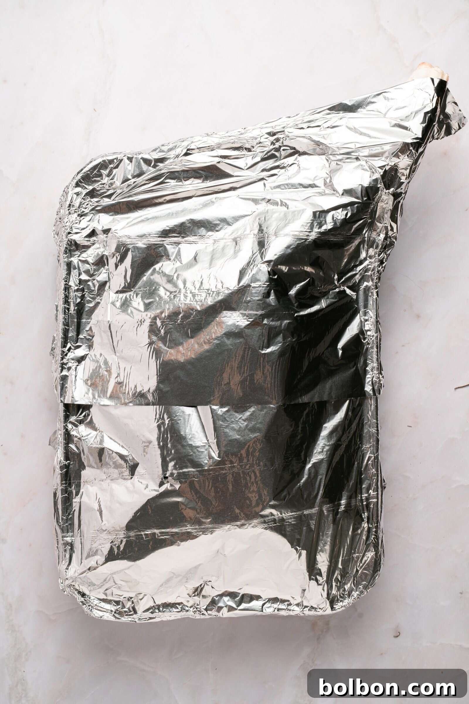 The Ultimate Roasted Lamb Leg 8 A rectangular baking dish covered tightly with aluminum foil sits on a light-colored marble surface.