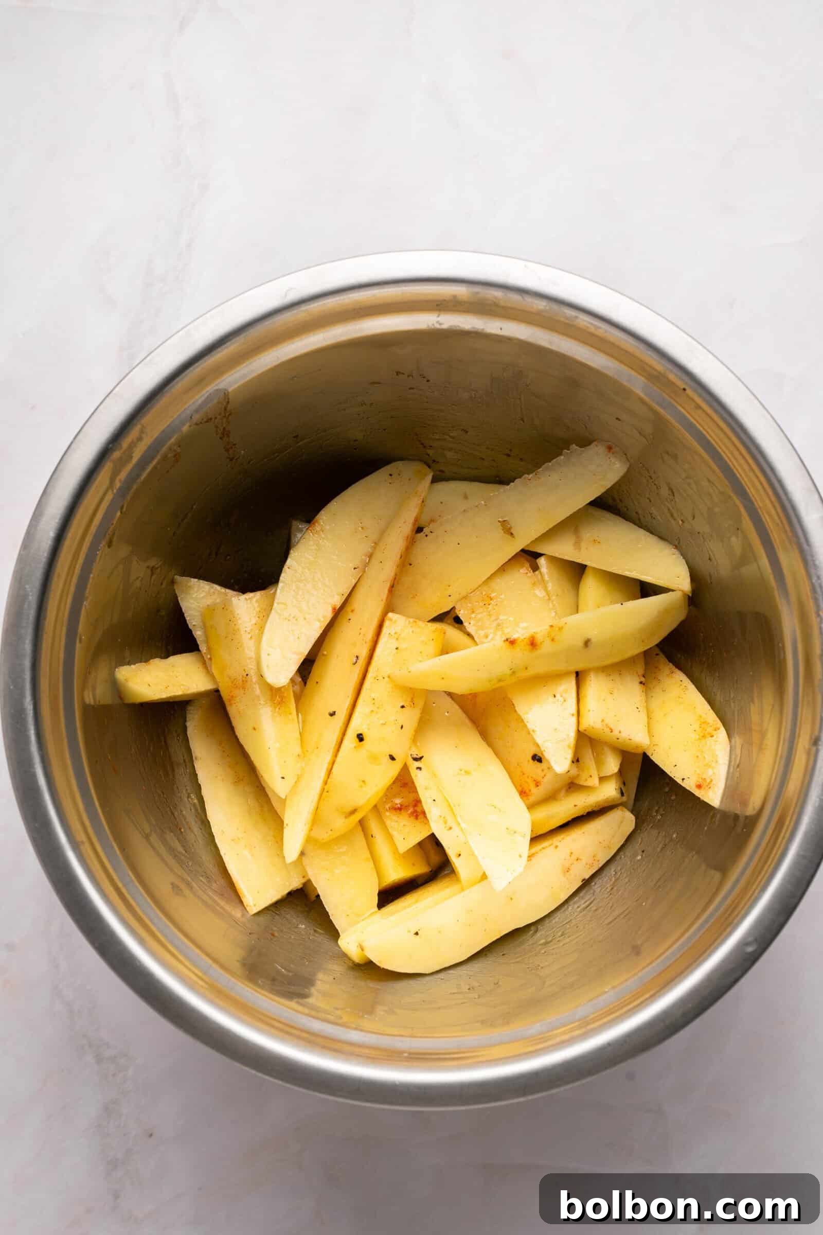 The Ultimate Roasted Lamb Leg 9 Raw potato wedges seasoned with spices in a stainless steel mixing bowl, placed on a light-colored countertop.