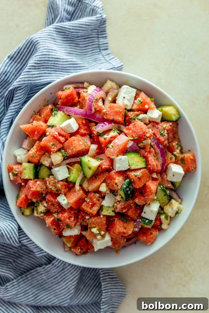 Sweet and Savory Watermelon Feta Salad 2 A vibrant Watermelon Feta Salad in a white bowl, garnished with fresh mint. Sliced red onions and crisp cucumbers are visible alongside the cubed watermelon and feta. A blue and white striped kitchen towel adds a touch of summer flair.