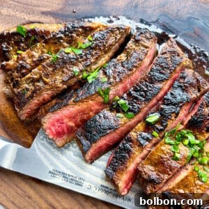 Sliced medium-rare steak with a seared crust, inspired by a classic Carne Asada recipe, garnished with chopped herbs and displayed on a wooden cutting board with a knife partially visible underneath.