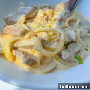 Bowl of creamy stew featuring a classic Liver and Onions Recipe, with tender meat slices, onions, and light sauce, garnished with chopped green herbs and served with a wooden spoon.