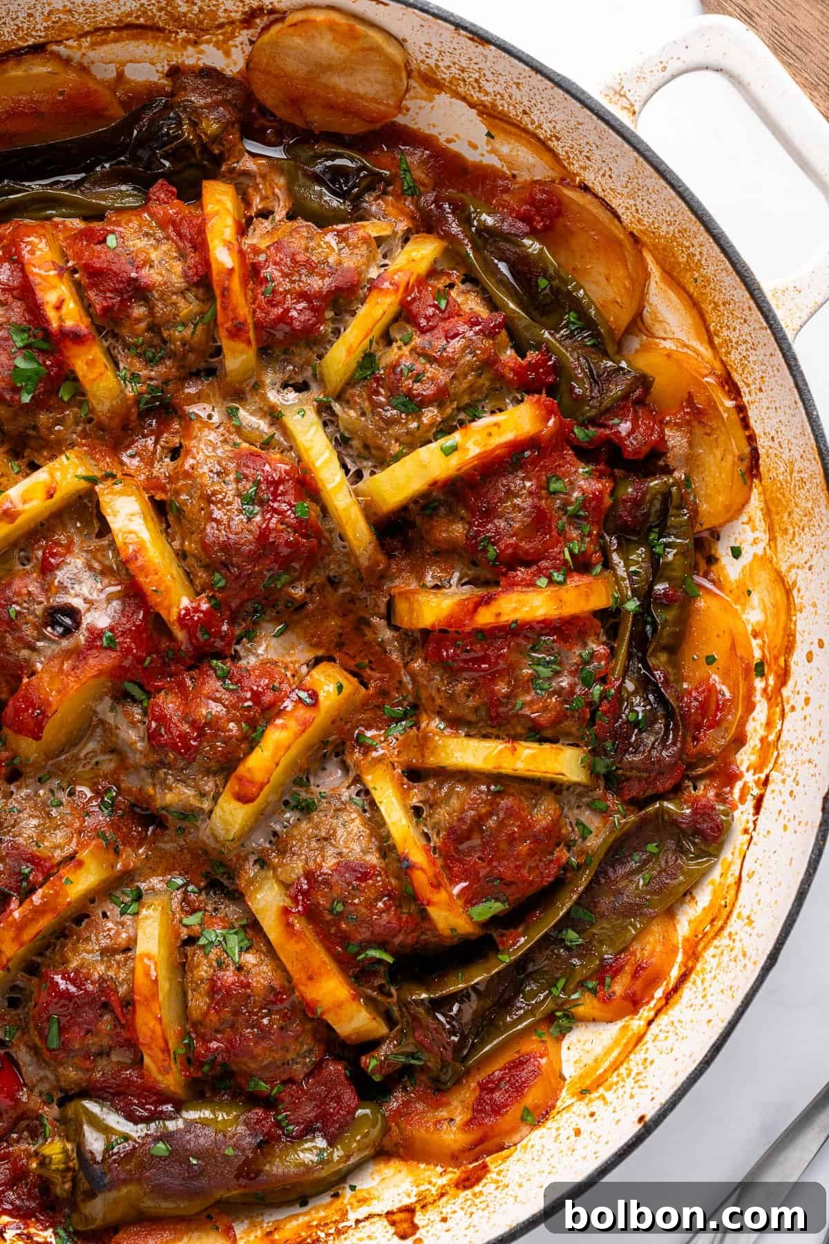 A close-up of a baked dish showing seasoned meatballs and potato wedges in a tomato-based sauce, garnished with chopped herbs. The dish is arranged in a circular pattern inside a white, round baking dish.