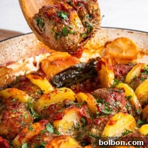 A wooden spoon holds a seasoned meatball from a dish with sliced potatoes, herbs, and bell peppers, all baked in a tomato sauce. The ingredients appear browned and garnished with fresh parsley.