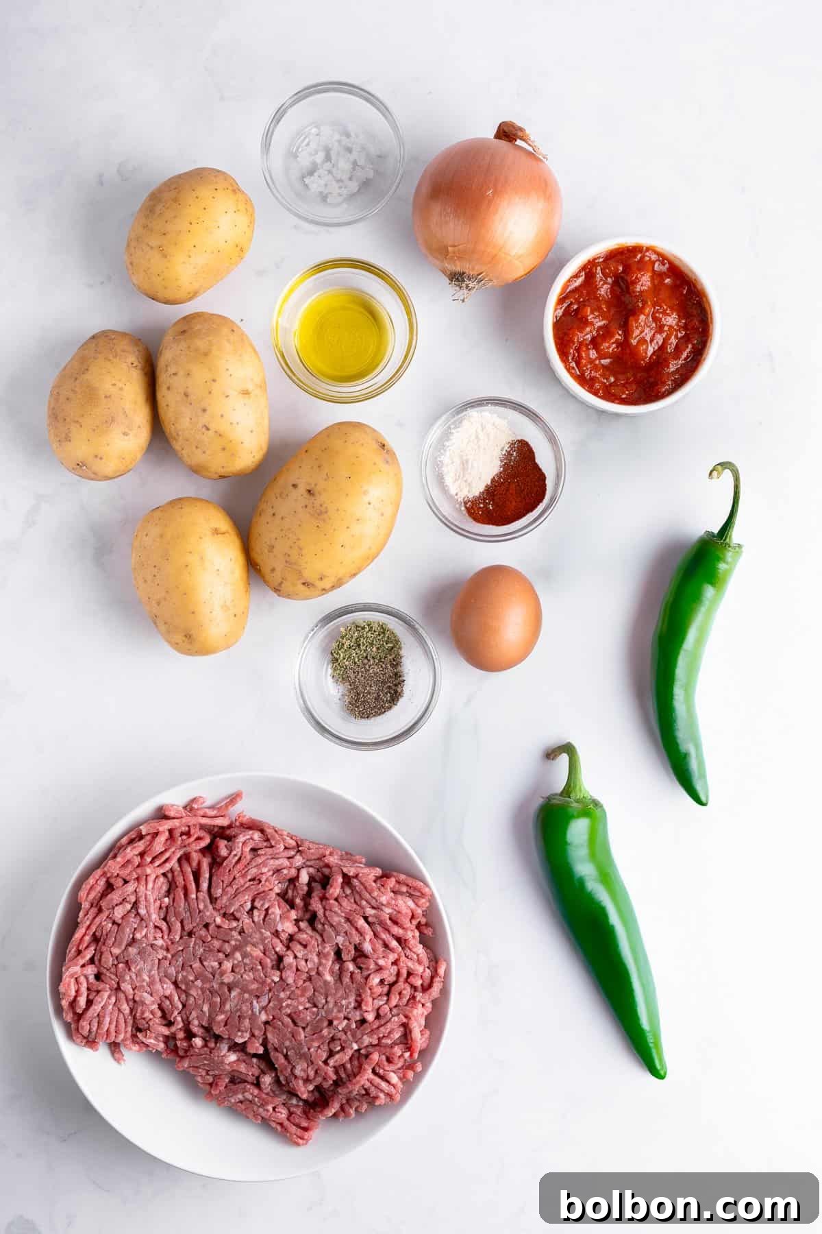 Flat lay of ingredients for a recipe: ground beef in a bowl, several potatoes, two green chili peppers, an onion, small bowls of tomato sauce, flour, spices, olive oil, salt, and a whole egg on a white surface.