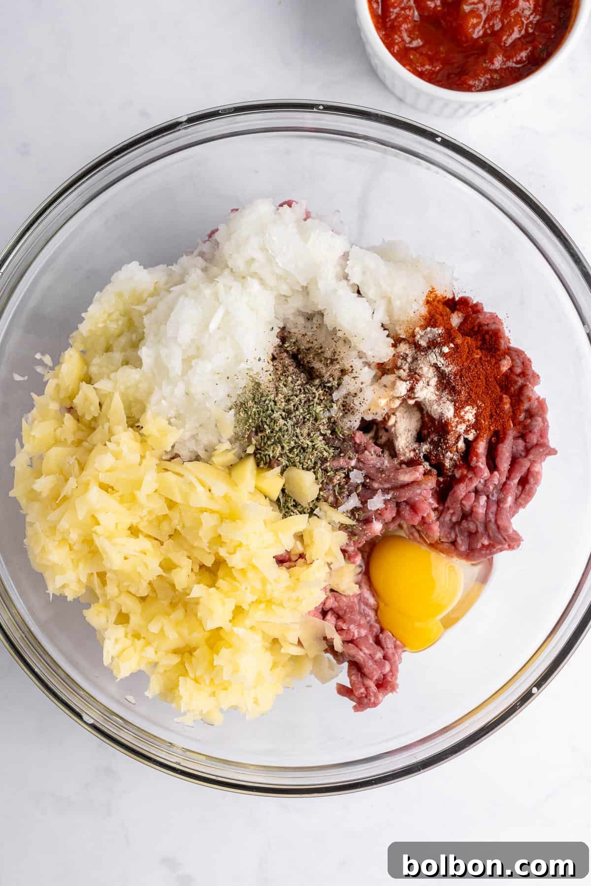 A glass bowl containing raw minced meat, chopped onions, potatoes, spices, and an egg yolk. A small bowl of red sauce is partially visible in the background on a white marble surface.