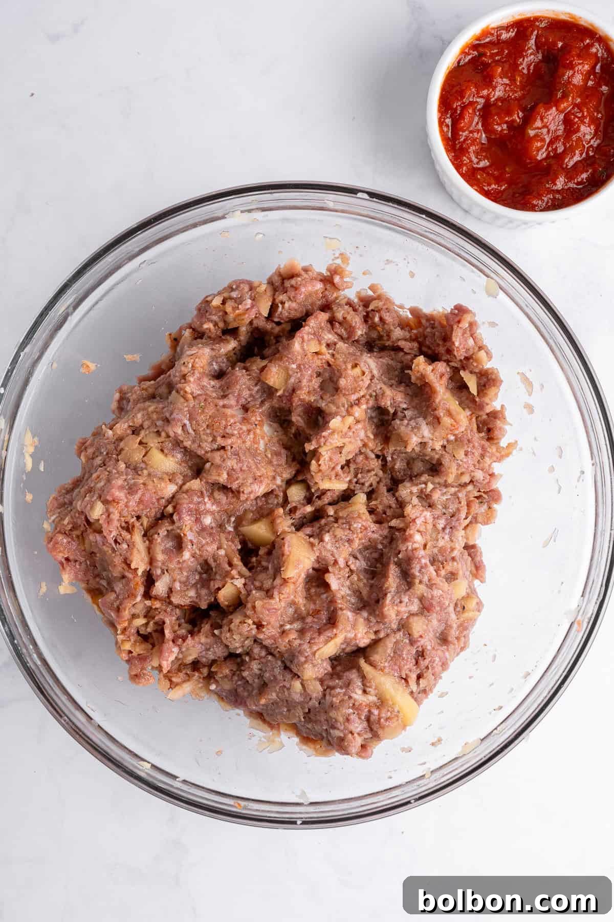 A glass bowl filled with seasoned minced meat on a marble counter. A small white bowl containing red sauce is positioned in the upper right corner.