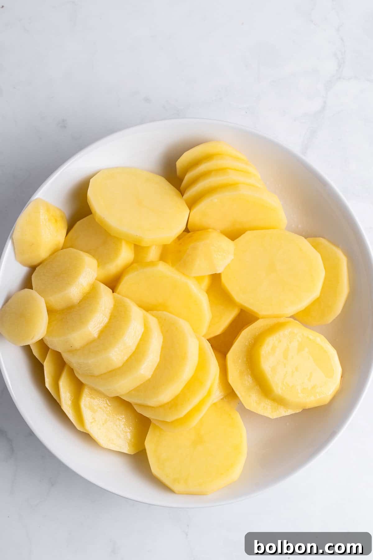 A white bowl filled with thin slices of yellow potatoes on a light marble surface.
