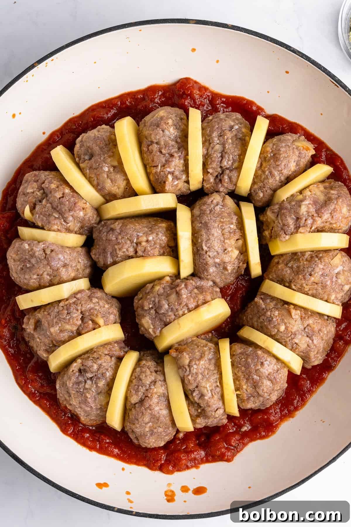 A round dish featuring a ring of uncooked meatloaf slices interspersed with potato slices, all placed over a thick layer of tomato sauce. The ingredients are neatly arranged, ready for cooking.
