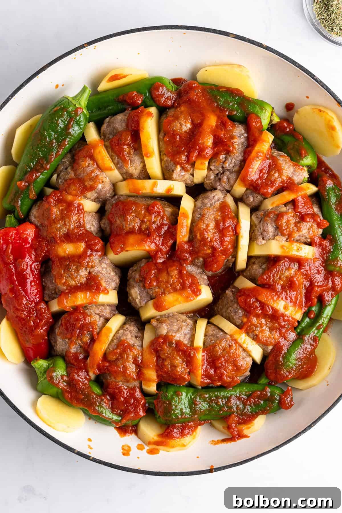 A round dish filled with sliced meat and potato layers, topped with tomato sauce. Surrounded by red and green peppers and additional potato slices. Garnished with herbs.