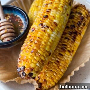 Three grilled ears of corn on the cob rest on parchment paper in a dish, with a small bowl of hot honey butter and a honey dipper beside them. The corn has a golden, lightly charred appearance—perfect Hot Honey Butter Corn.