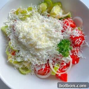 A white bowl filled with sliced tomatoes, cucumbers, onions, and peppers is topped with a generous amount of grated white cheese and parsley—classic ingredients of a traditional Shopska Salad.