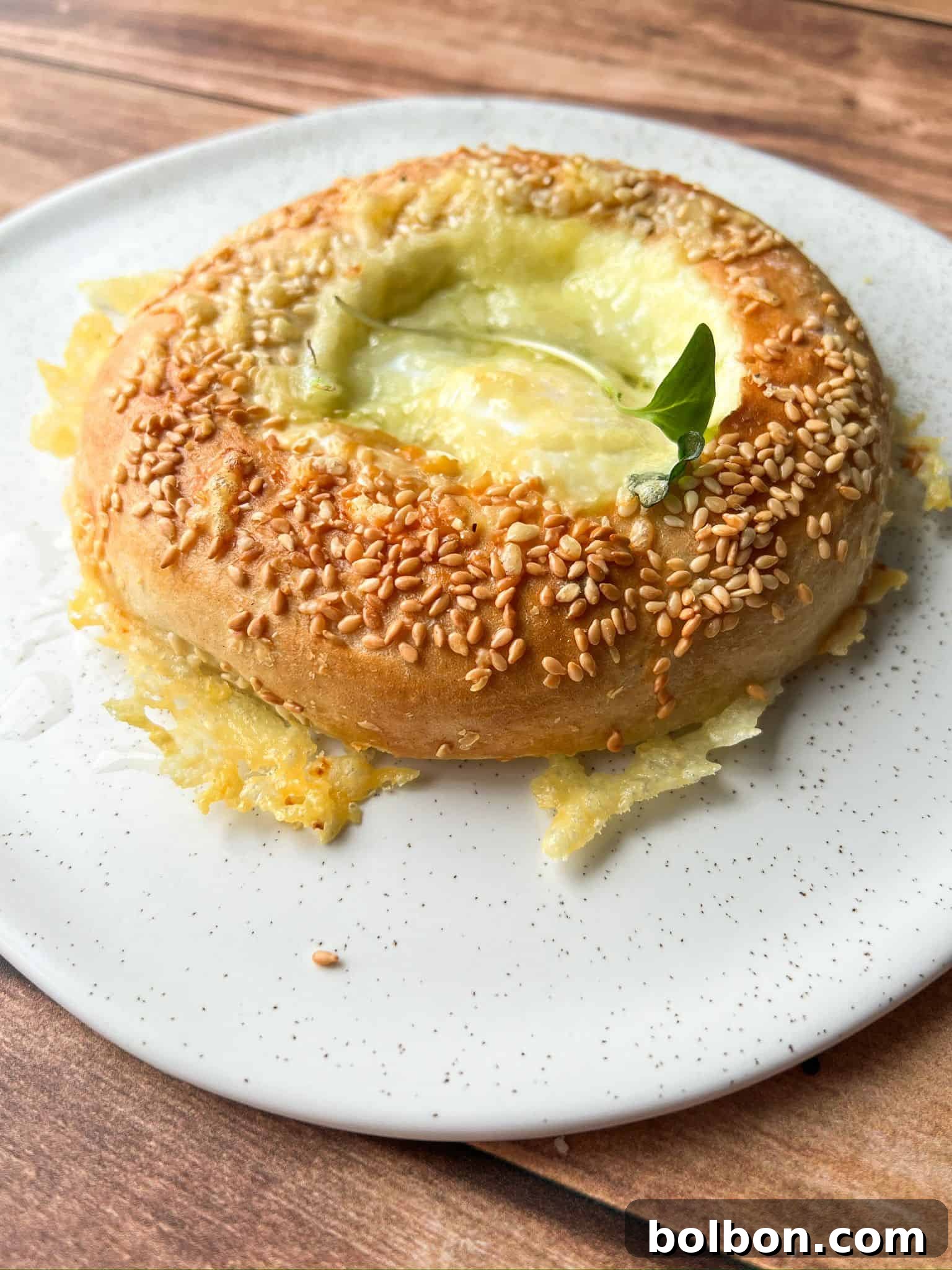 A round, baked bread topped with sesame seeds and melted cheese, garnished with a small green leaf, sits on a white speckled plate on a wooden surface.
