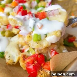 A close-up of a fork lifting loaded cauliflower nachos topped with diced red tomatoes, green onions, red onions, melted cheese, and a drizzle of white sauce on a parchment-lined tray.