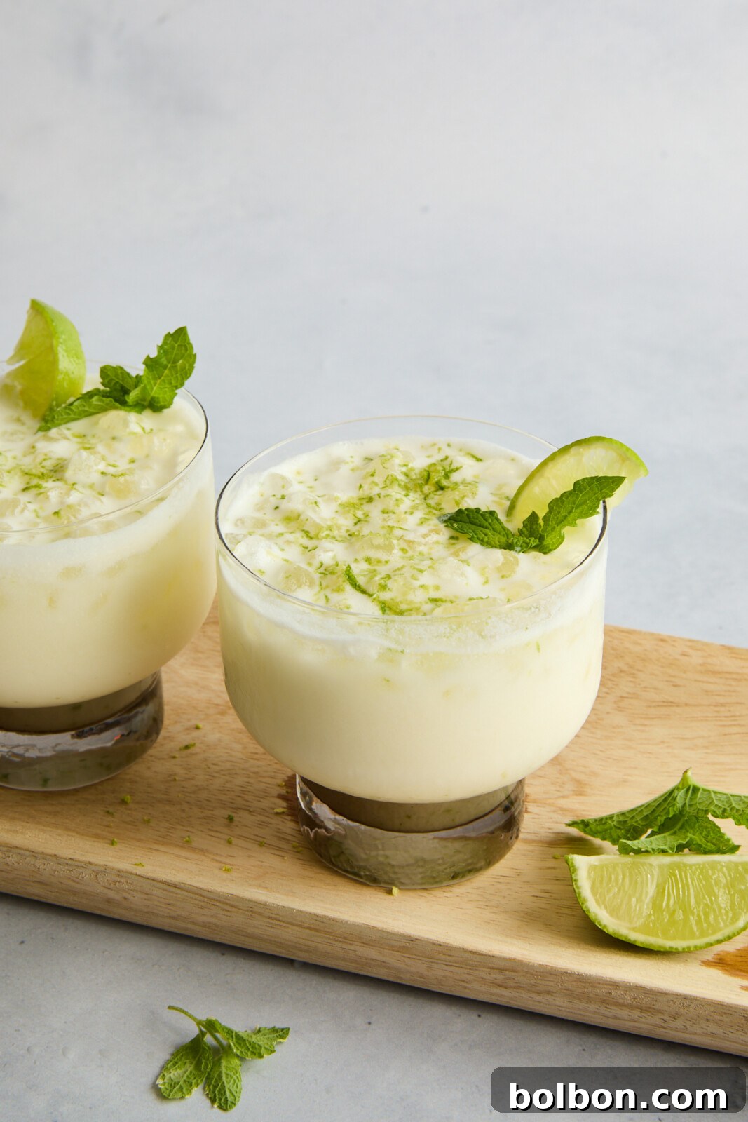 Brazilian Lemonade served in short, transparent glasses on a wooden board. Each drink is garnished with a lime wedge and a sprig of mint. A pile of lime zest and mint leaves are sprinkled on top for decoration, with extra lime wedges on the board.