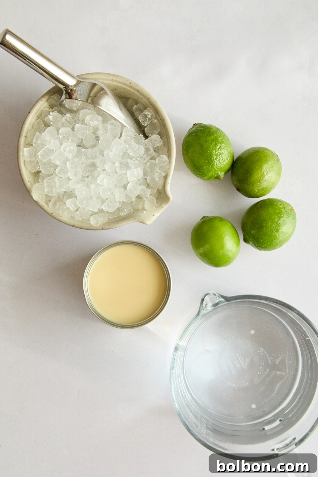 Top view of ingredients for a lime ice drink on a white surface. Includes a container of crushed ice with a metal scoop, three whole green limes, an open can of condensed milk, and a glass jug filled with clear liquid.