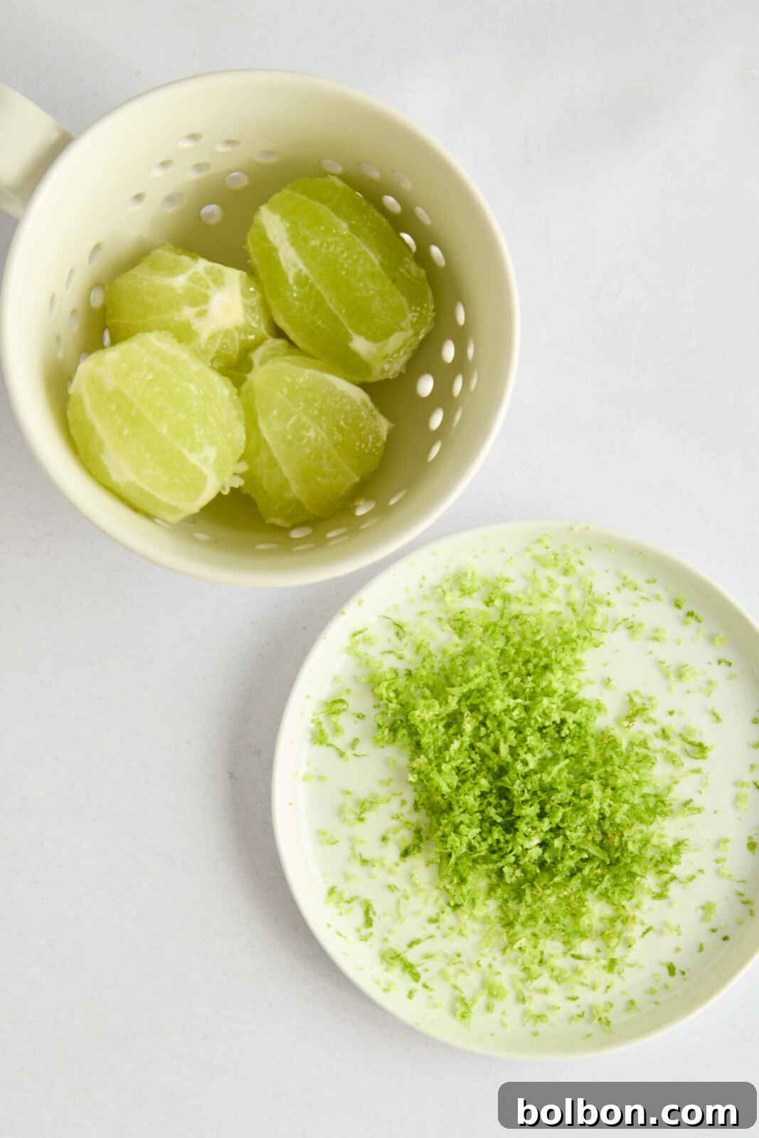 A white bowl with peeled lime segments rests next to a white plate containing a pile of green lime zest. Both items are placed on a white surface.