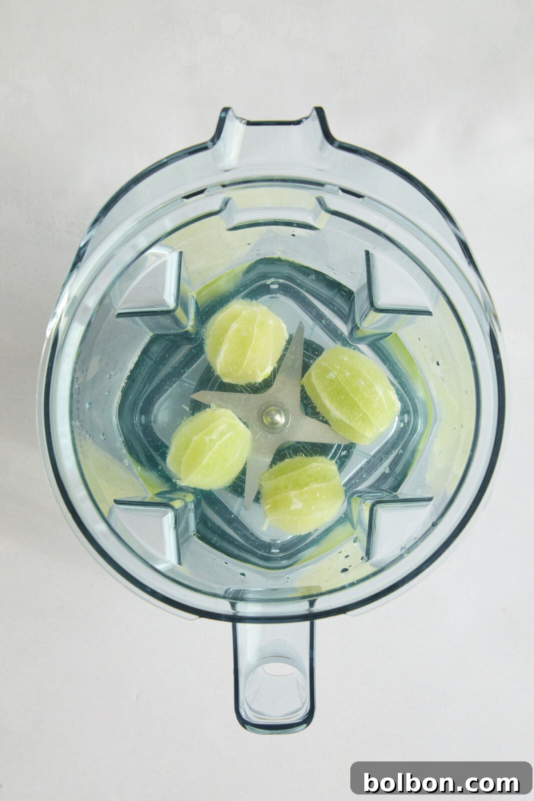 Top view of a blender jug containing four roughly chopped limes placed around the central metal blades. The blender jug is transparent, showing the limes and some small bits of skin against the plain white background.