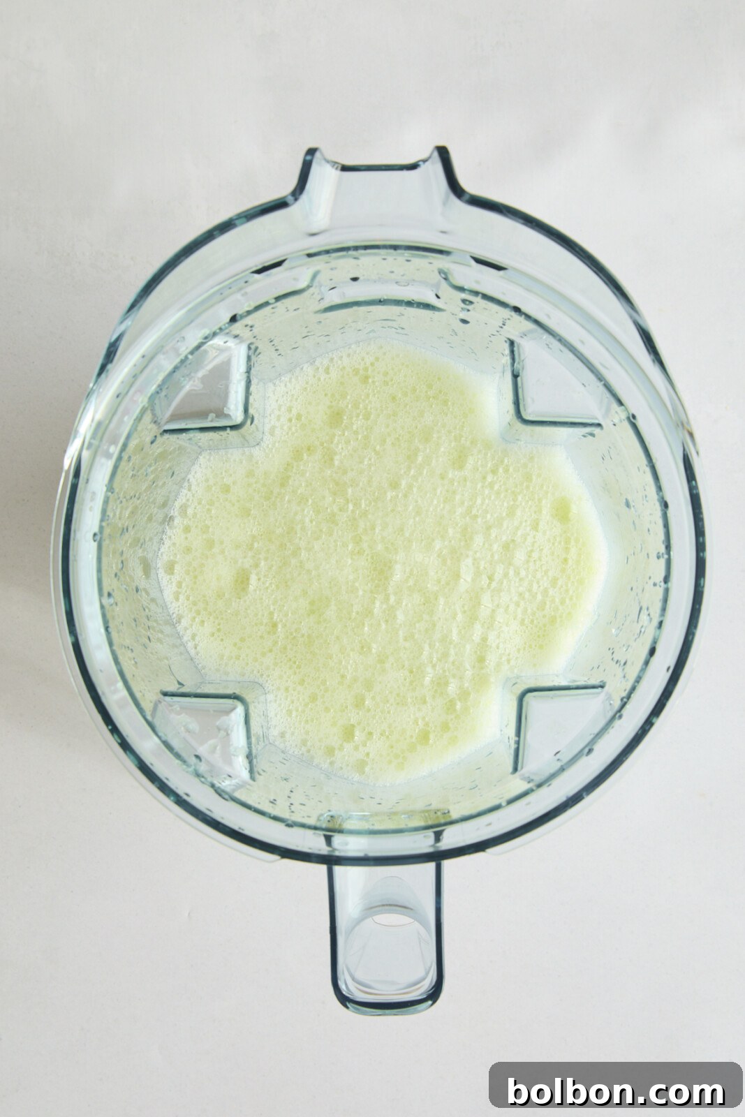 A top-down view of a blender filled with a light green, frothy Brazilian lemonade mixture. The blender is placed on a light-colored surface, and four prongs are visible inside the container, which is about halfway full of the blended liquid.