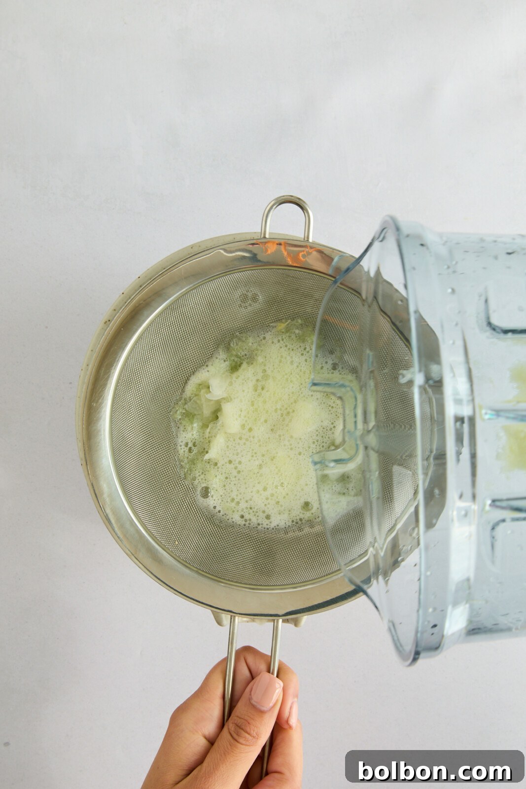 A hand holds a mesh strainer over a bowl while pouring blended celery juice from a blender jug into the strainer. The image captures the juice being strained, with some pulp accumulated in the strainer.