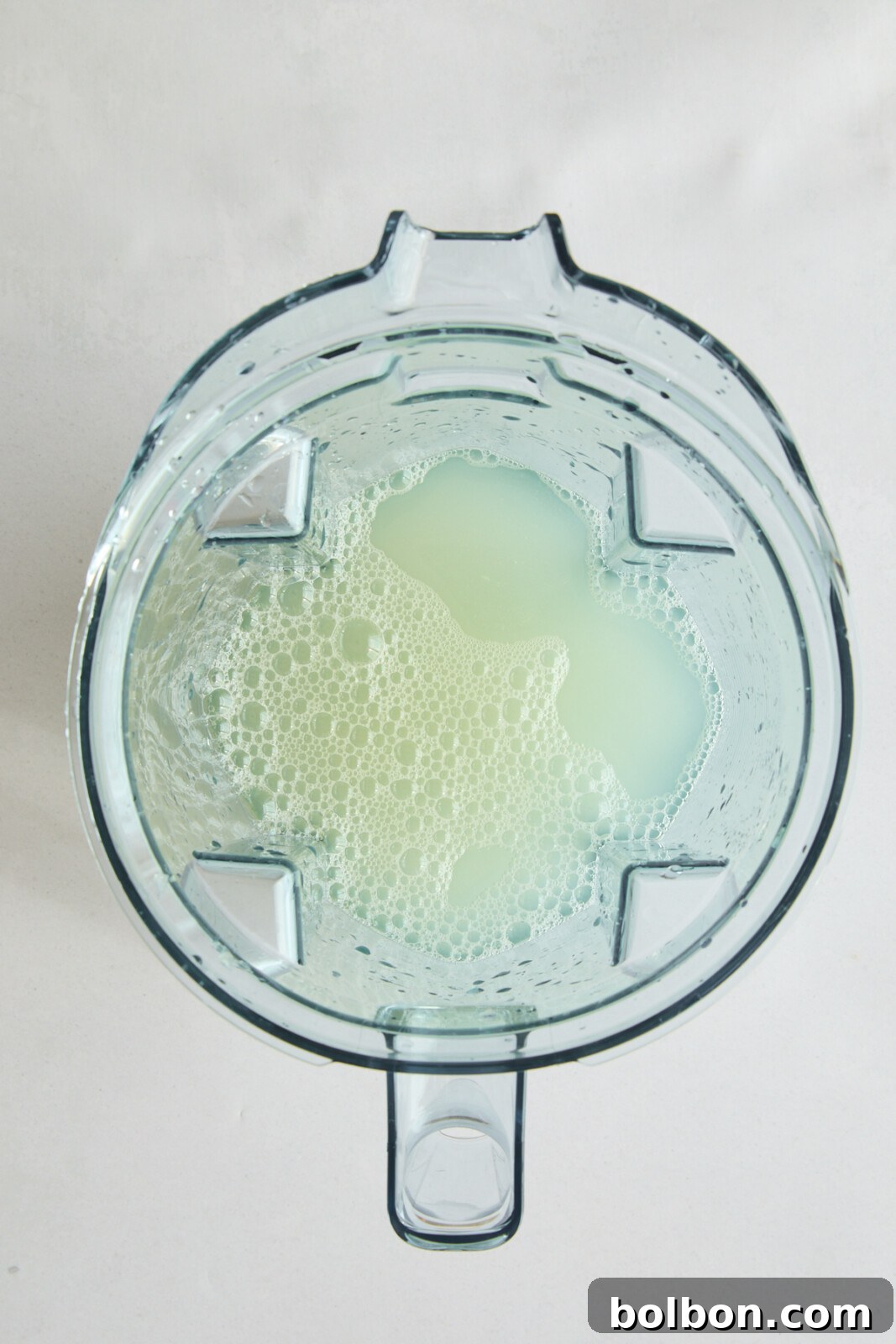 Top view of a blender pitcher containing a light green, frothy liquid. The inside of the blender is clear, showing the creamy texture and bubbles formed on the surface of the mixture against a white background.