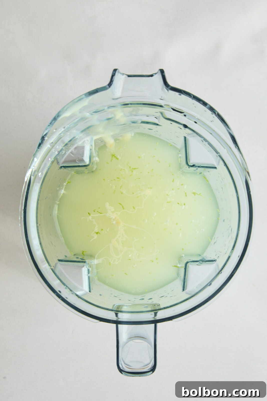 Top view of a blender containing a pale green, partially blended liquid with small bits of pulpy and fibrous material. The blender is placed on a light, almost white surface.