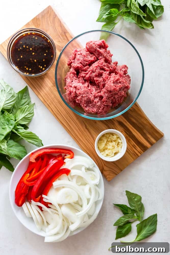 Spicy Thai Basil Beef 3 Ingredients for Thai basil beef - a bowl with the sauce, ground beef, minced garlic, red bell peppers and onions, surrounded by fresh basil leaves on a wooden board.