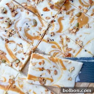A close-up of Cottage Cheese Bark dessert bars with a creamy white topping swirled with caramel and sprinkled with granola, sitting on parchment paper. A knife is partially placed under one of the bars.