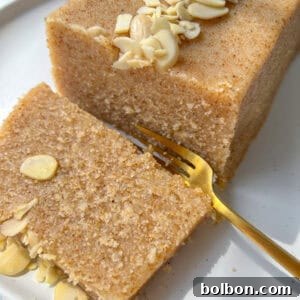 A close-up of a rectangular slice of Semolina Cake topped with sliced almonds on a white plate. A gold fork rests beside the cake, with one piece of the Semolina Cake partially cut and ready to eat.