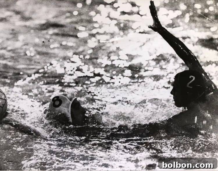 Black and white photo of a water polo match. Two players are shown in the pool, one wearing a cap with the number 8 and another with the number 2. The player with number 2 has their arm raised, with water splashing around them.