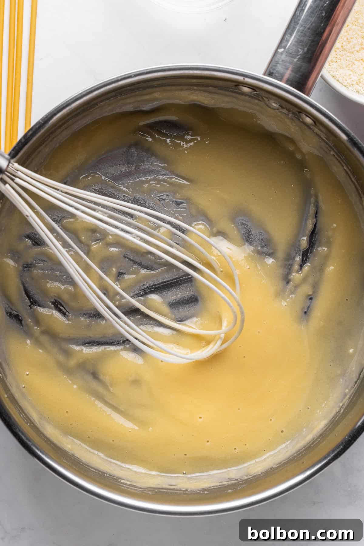 A stainless steel bowl with creamy yellow batter being mixed with a whisk. Spaghetti and a metal pot with breadcrumbs are nearby on a white countertop.