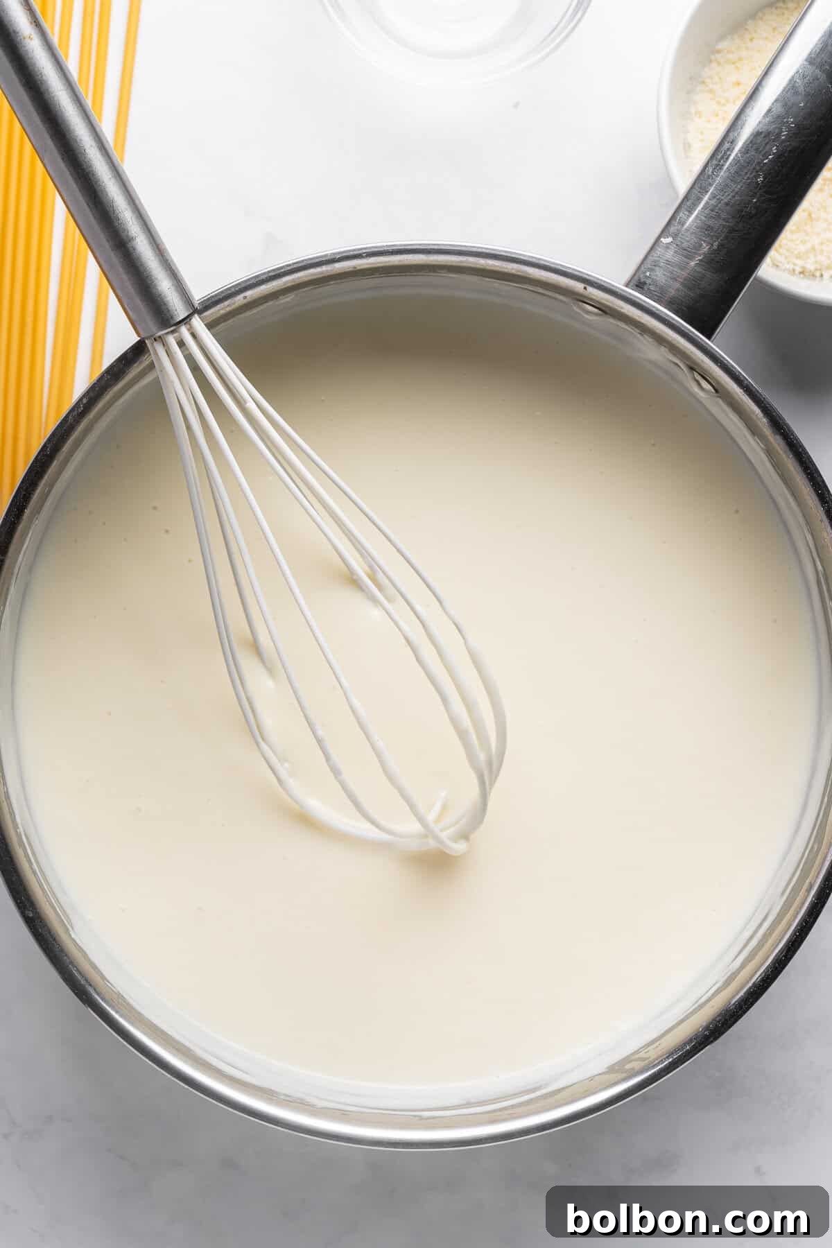 A whisk in a pot of creamy white sauce, likely béchamel, on a light countertop. Uncooked spaghetti and a bowl of breadcrumbs are partially visible nearby.