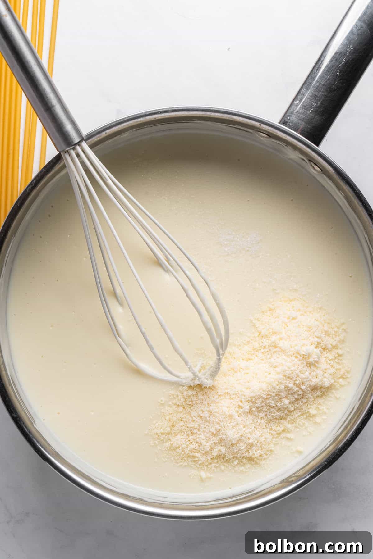 A saucepan filled with white sauce being whisked, with a pile of grated cheese on top for the Pastitsio recipe. Uncooked spaghetti is visible in the background on the countertop.