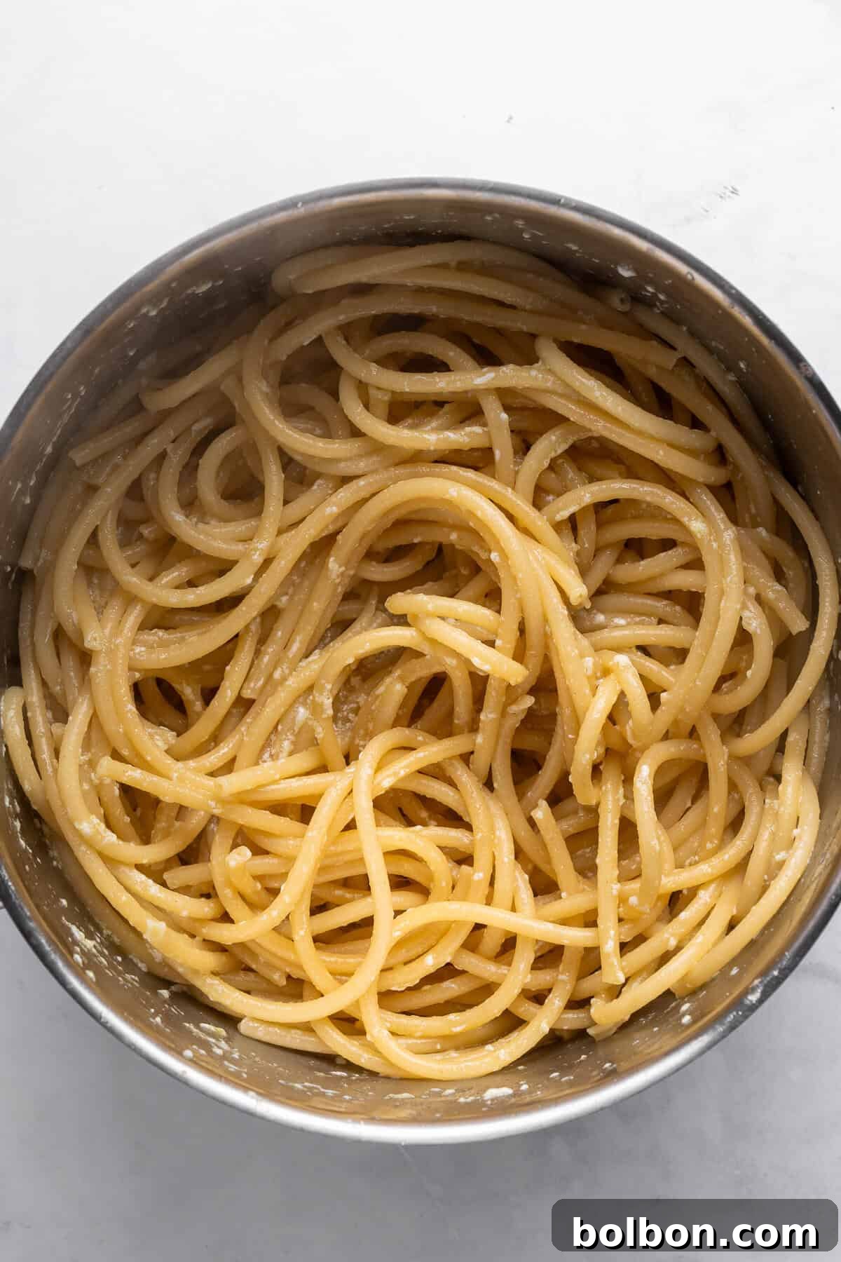 A stainless steel pot filled with spaghetti lightly coated in a creamy parmesan sauce, set on a white marble surface. The pasta is neatly arranged, highlighting the cheese glistening on the strands.