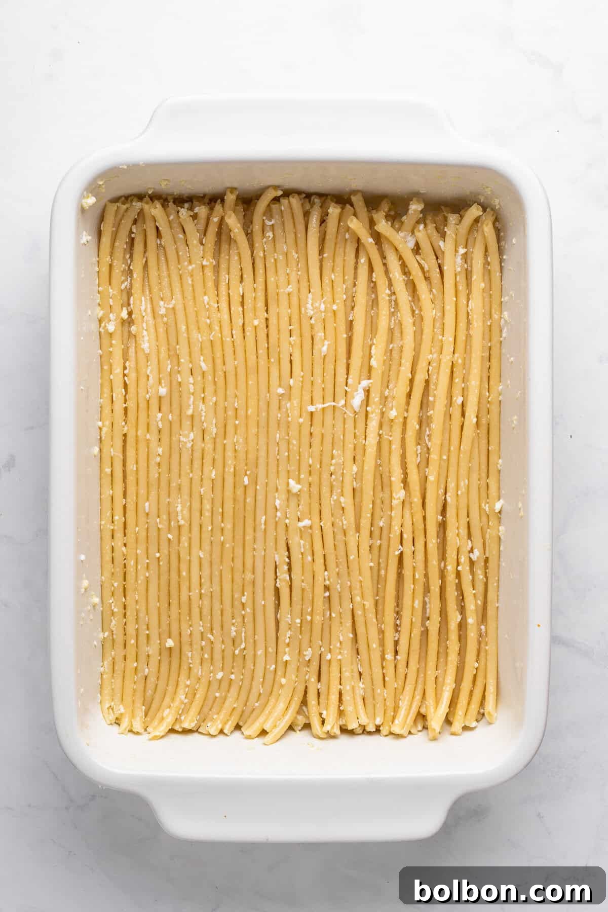 A baking dish filled with neatly arranged, uncooked pasta noodles topped with grated cheese on a white marble countertop.