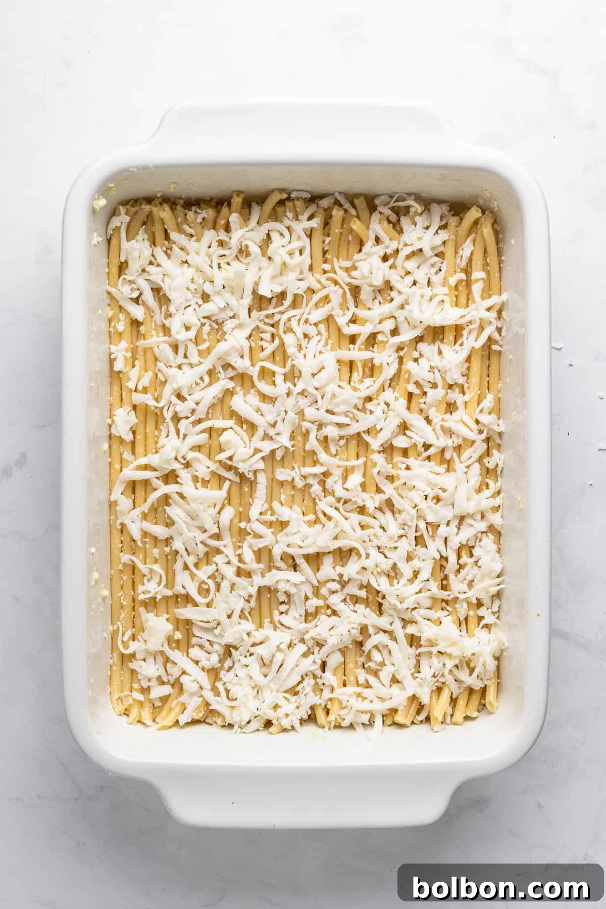 A white rectangular baking dish filled with lasagna noodles covered in shredded cheese, ready for baking. The background is a marble countertop.