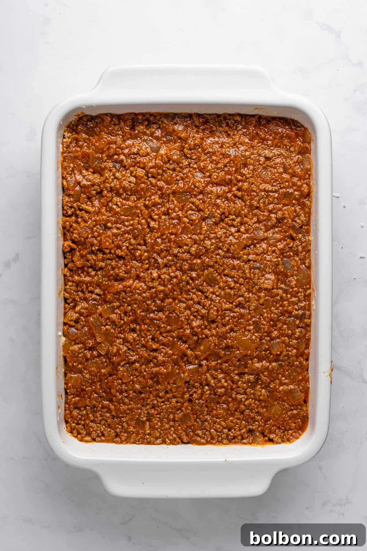 A white rectangular baking dish filled with a layer of finely ground meat sauce, cooked to a rich brown color, is placed on a light gray marble surface.