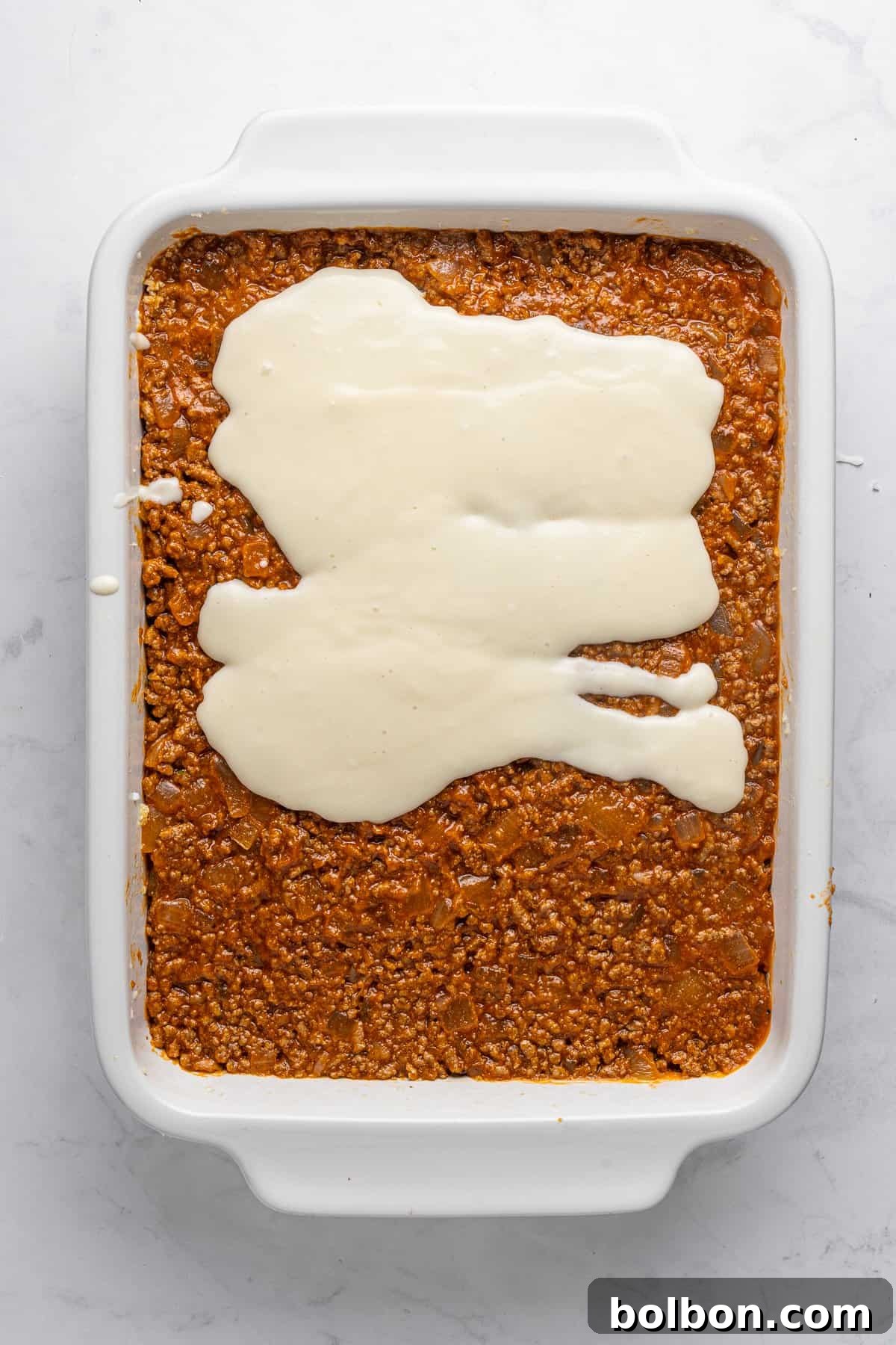 A rectangular white baking dish filled with a layer of cooked minced meat topped with a light tan sauce, spread unevenly. The dish sits on a marbled white countertop.
