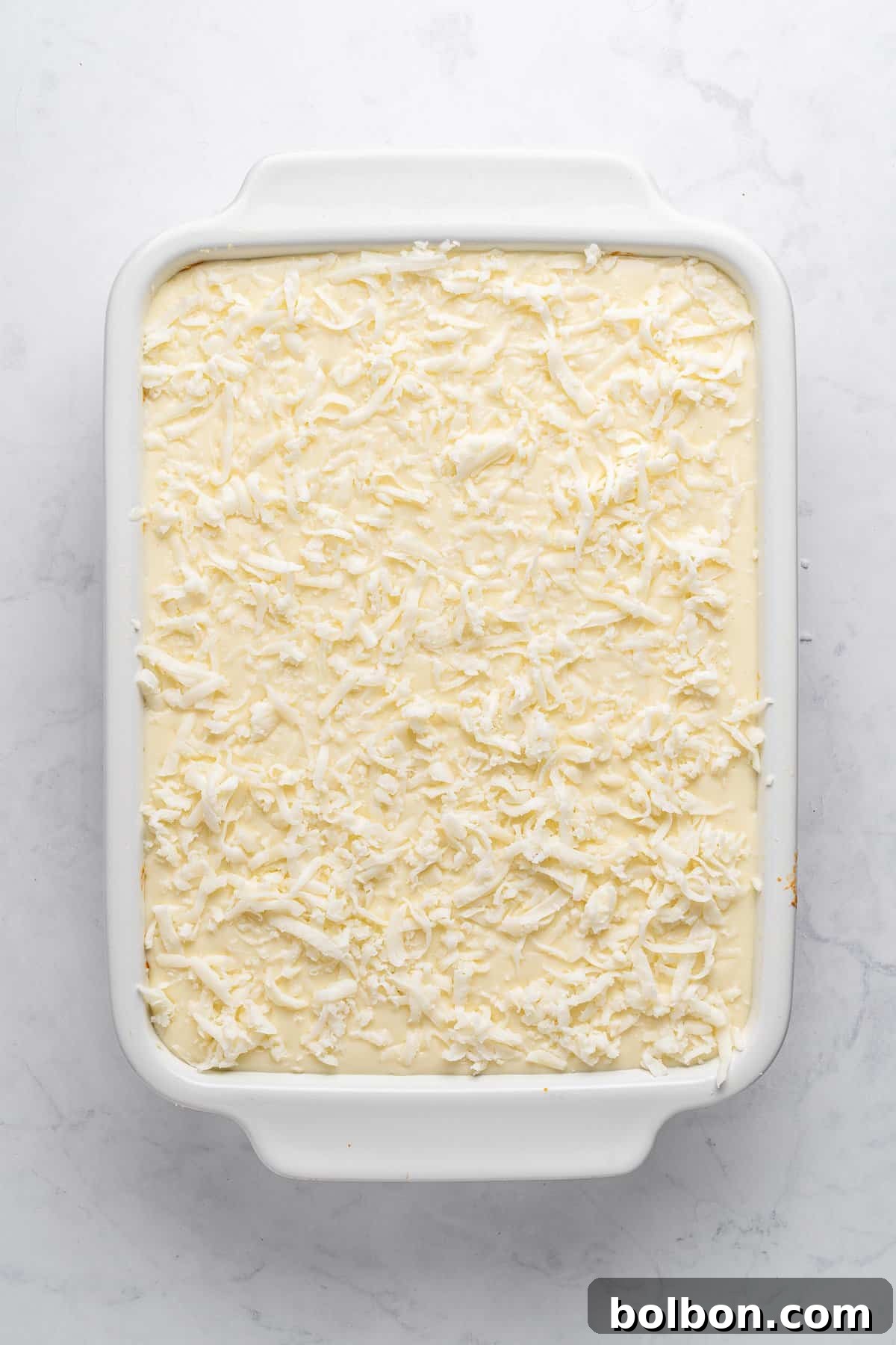 A rectangular white baking dish filled with a Pastitsio recipe topped with a layer of shredded coconut. The dish is set against a light gray marble surface.