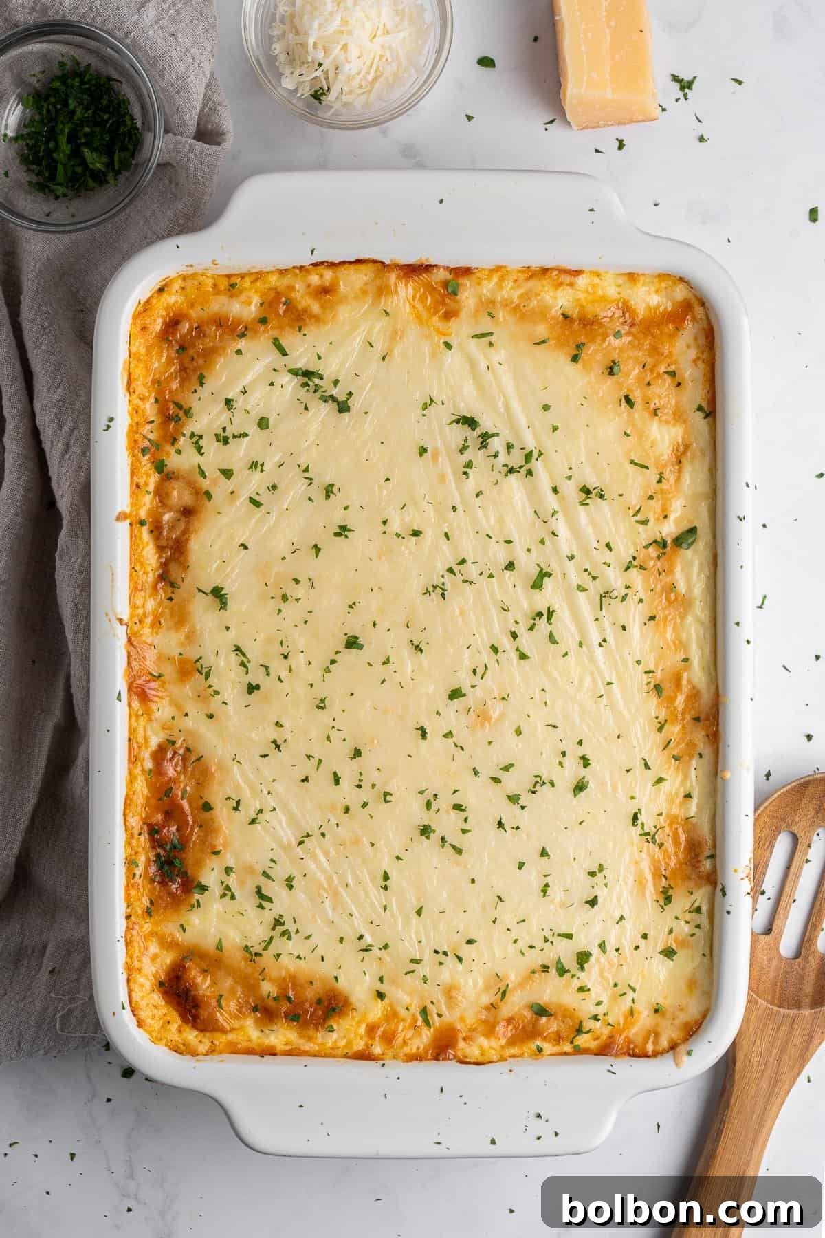 A golden-brown baked Pastitsio recipe in a white rectangular dish, garnished with chopped parsley. Surrounding the dish are bowls of grated cheese and chopped herbs, a block of cheese, and a wooden spatula on a light-colored countertop.