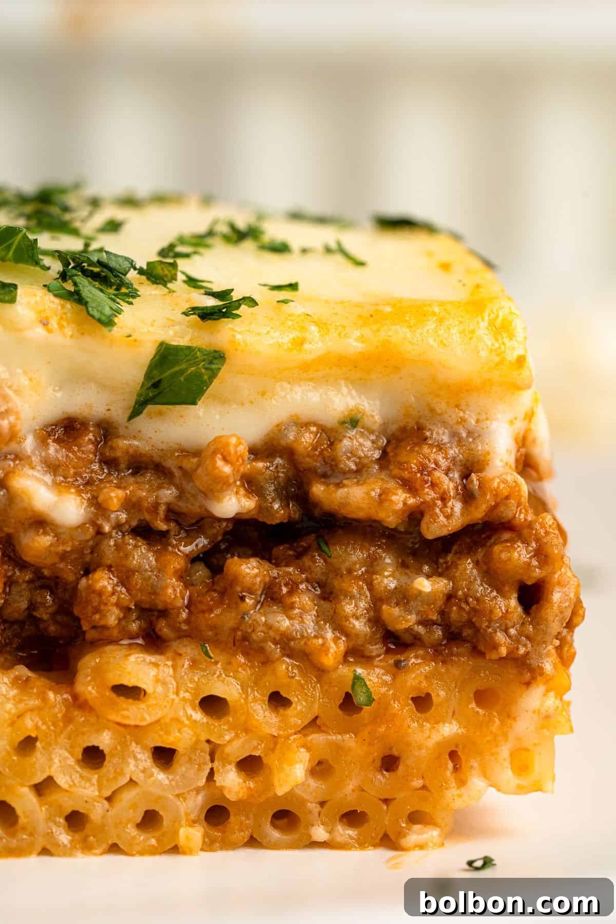 A close-up of a slice of pastitsio, featuring layers of tubular pasta, savory meat sauce, and creamy béchamel on top. Garnished with chopped parsley.