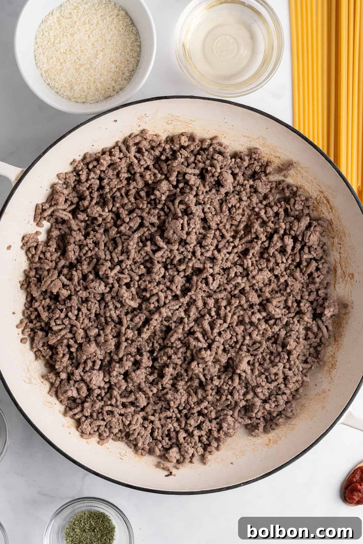 A pan filled with cooked ground meat sits on a countertop. Nearby, uncooked spaghetti, a bowl of breadcrumbs, a small glass bowl of oil, and various spices are positioned, ready for meal preparation.