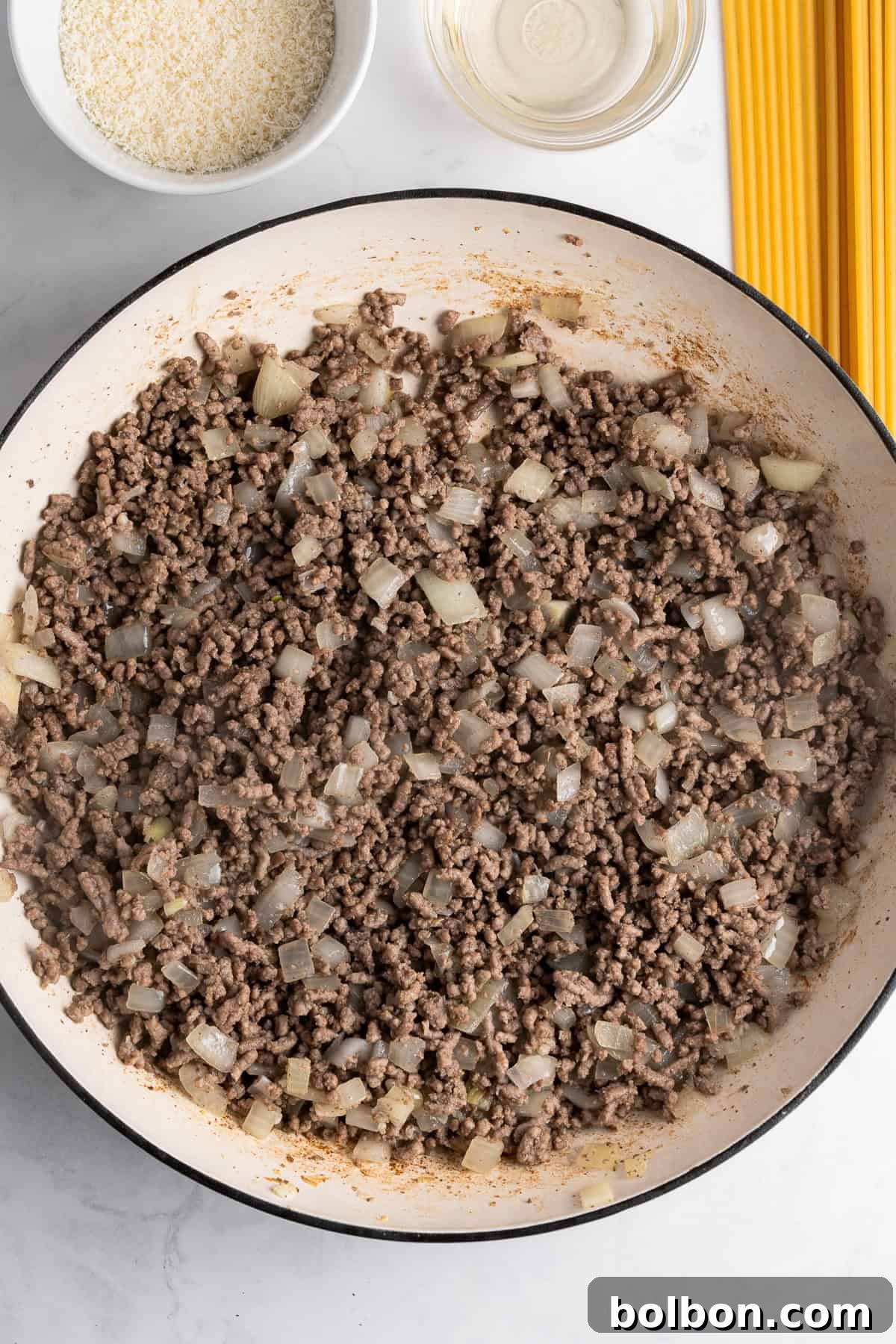 A large pan contains cooked ground beef mixed with diced onions. Nearby are uncooked spaghetti strands, a small bowl of grated cheese, and a glass bowl of clear liquid on a light marble surface.