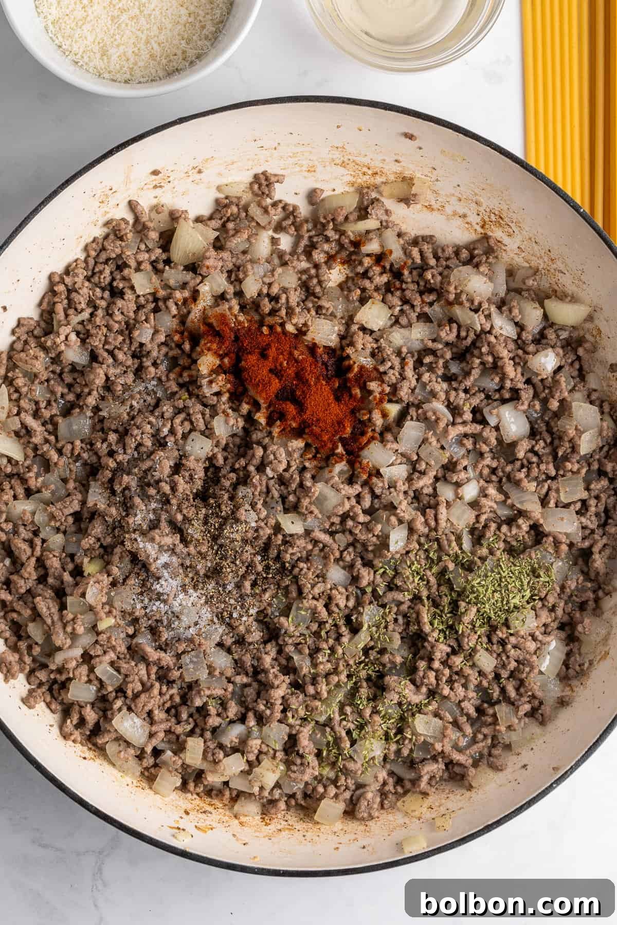 A pan filled with cooked ground beef, chopped onions, and a mix of spices including paprika, black pepper, salt, and herbs. Beside the pan are a bowl of grated cheese, a clear liquid, and uncooked spaghetti on a marble countertop.