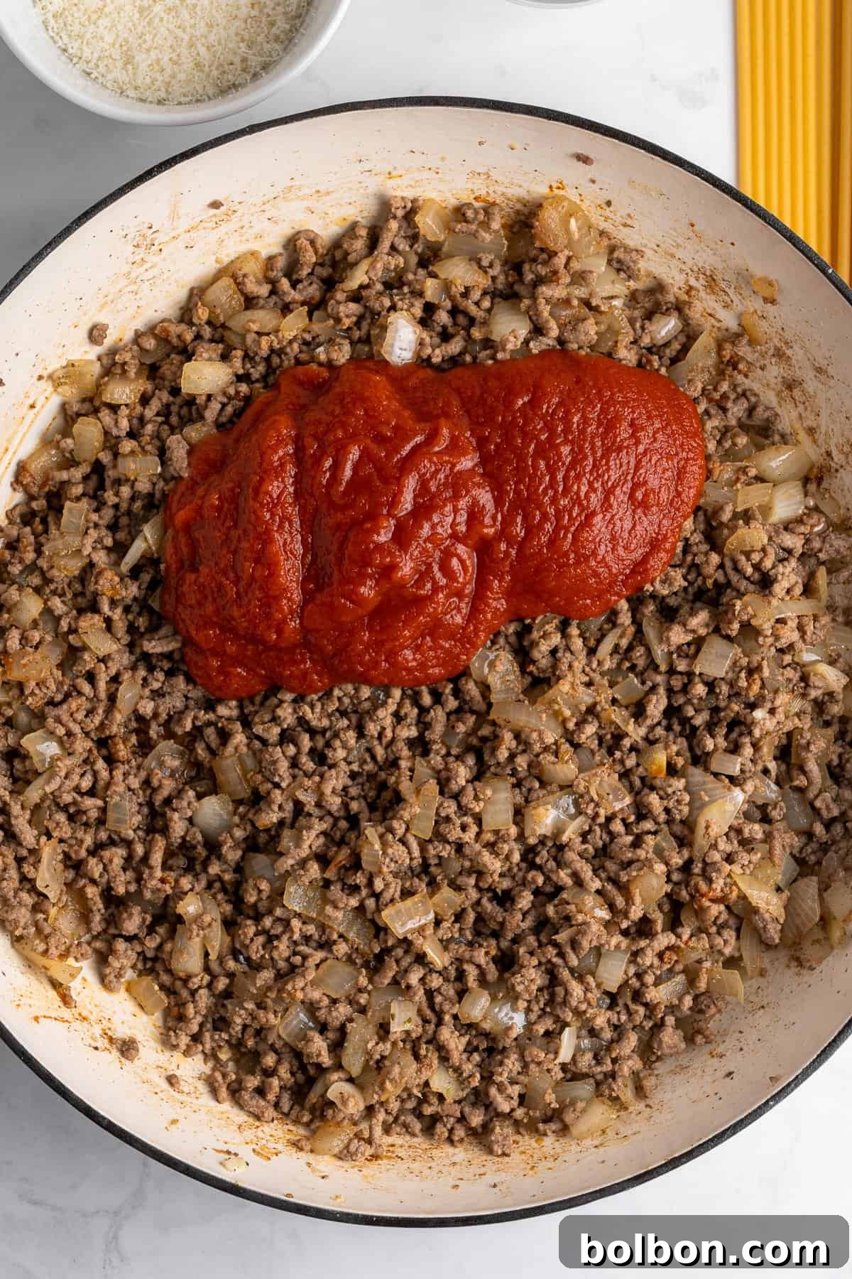 A skillet containing cooked ground beef and diced onions, topped with a generous amount of tomato paste. In the background, there's a bowl of grated cheese and uncooked spaghetti on a marble surface.