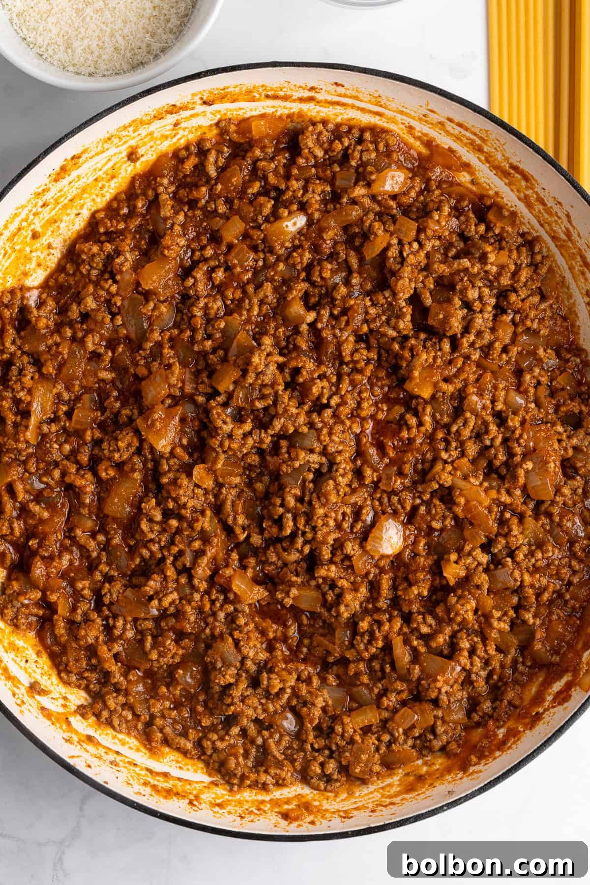 A close-up of a pan filled with rich red meat sauce, containing ground meat and visible bits of onion, with a small plate of grated cheese and uncooked spaghetti nearby. The sauce appears thick and well-cooked.