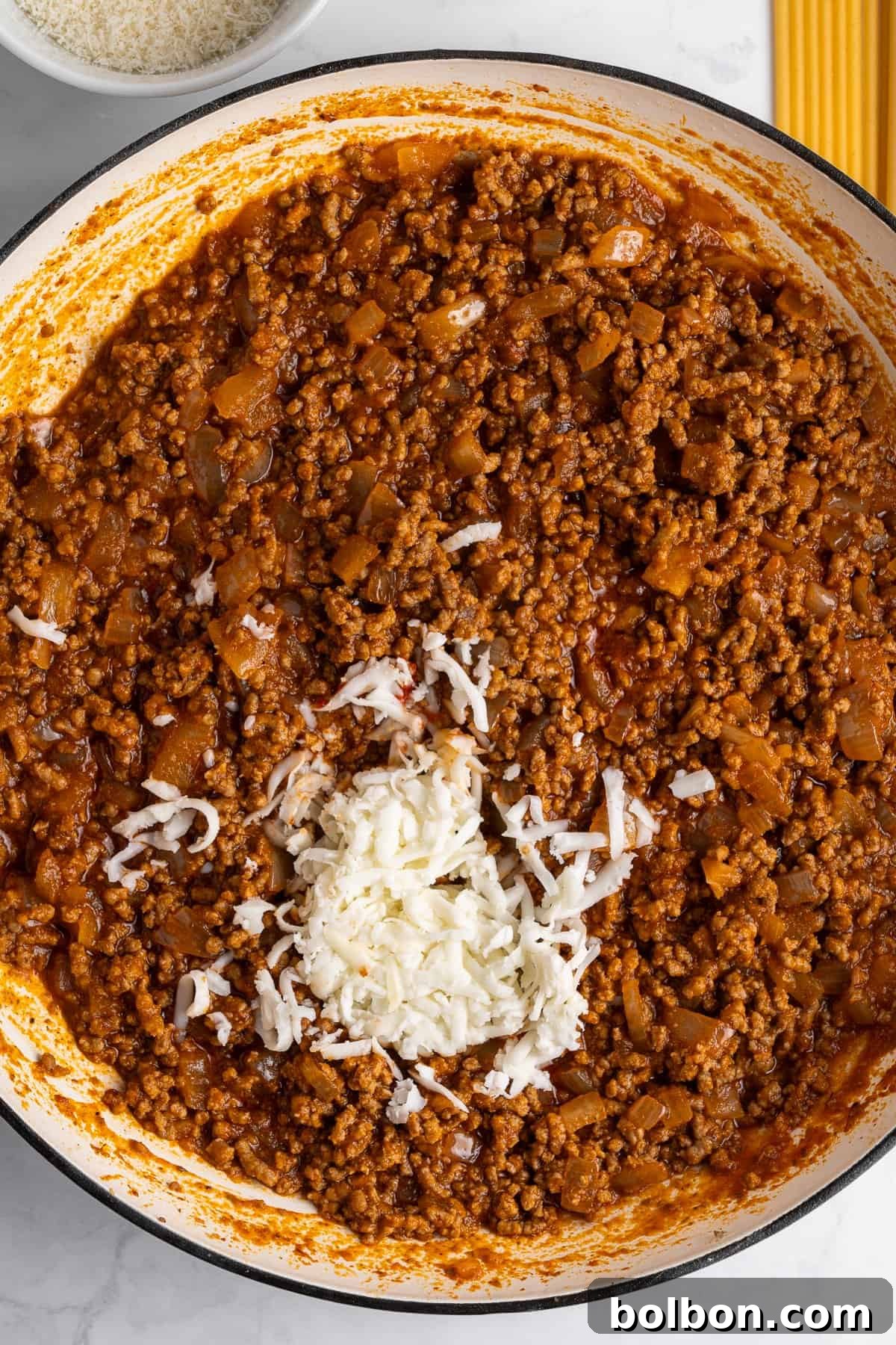A pot filled with savory bolognese sauce made from ground meat and diced vegetables for the Pastitsio recipe. There's a pile of grated cheese melting in the center. Uncooked spaghetti and a bowl of grated cheese on the side.